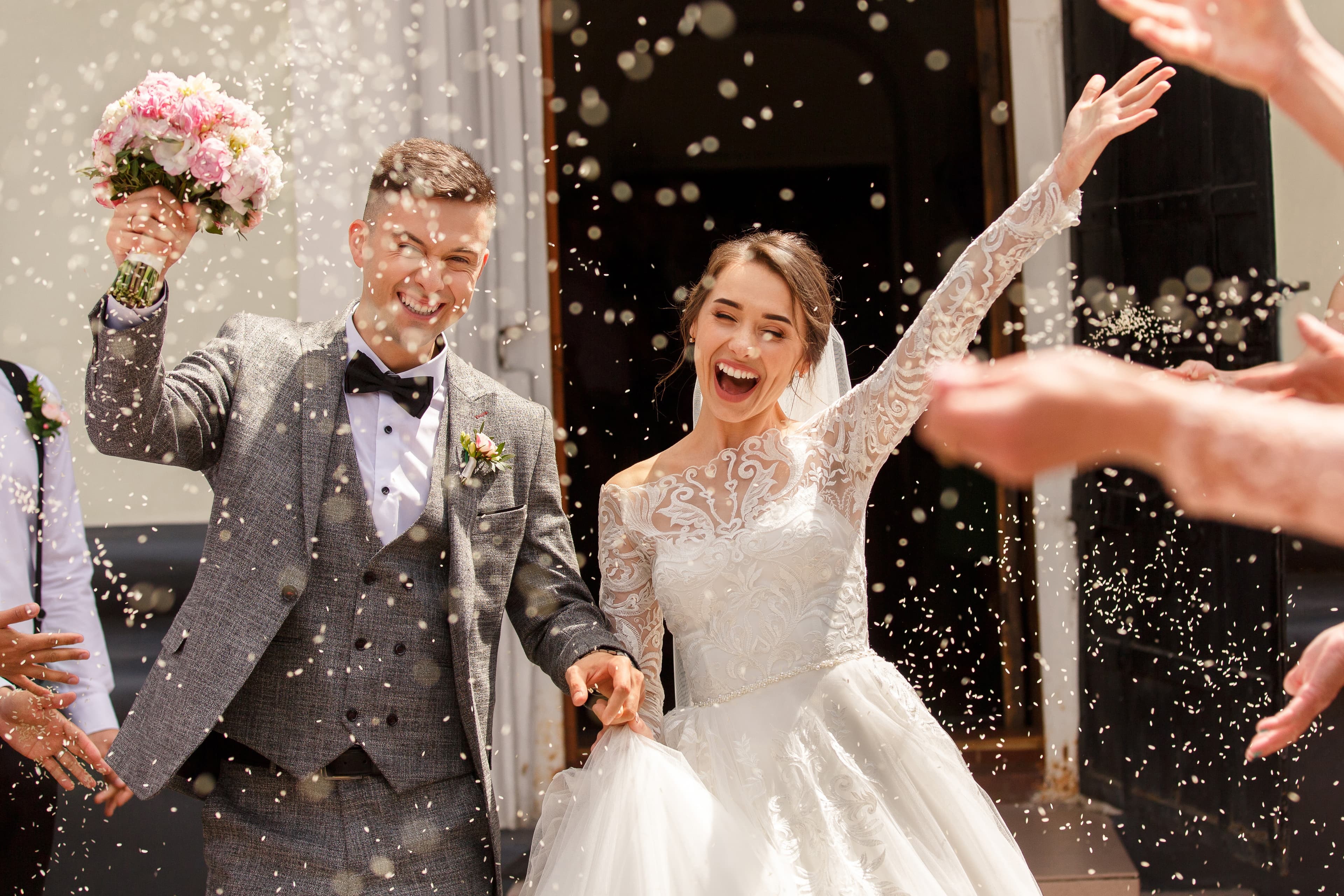 A smiling bride and groom in festive attire leave a church while being showered with rice.