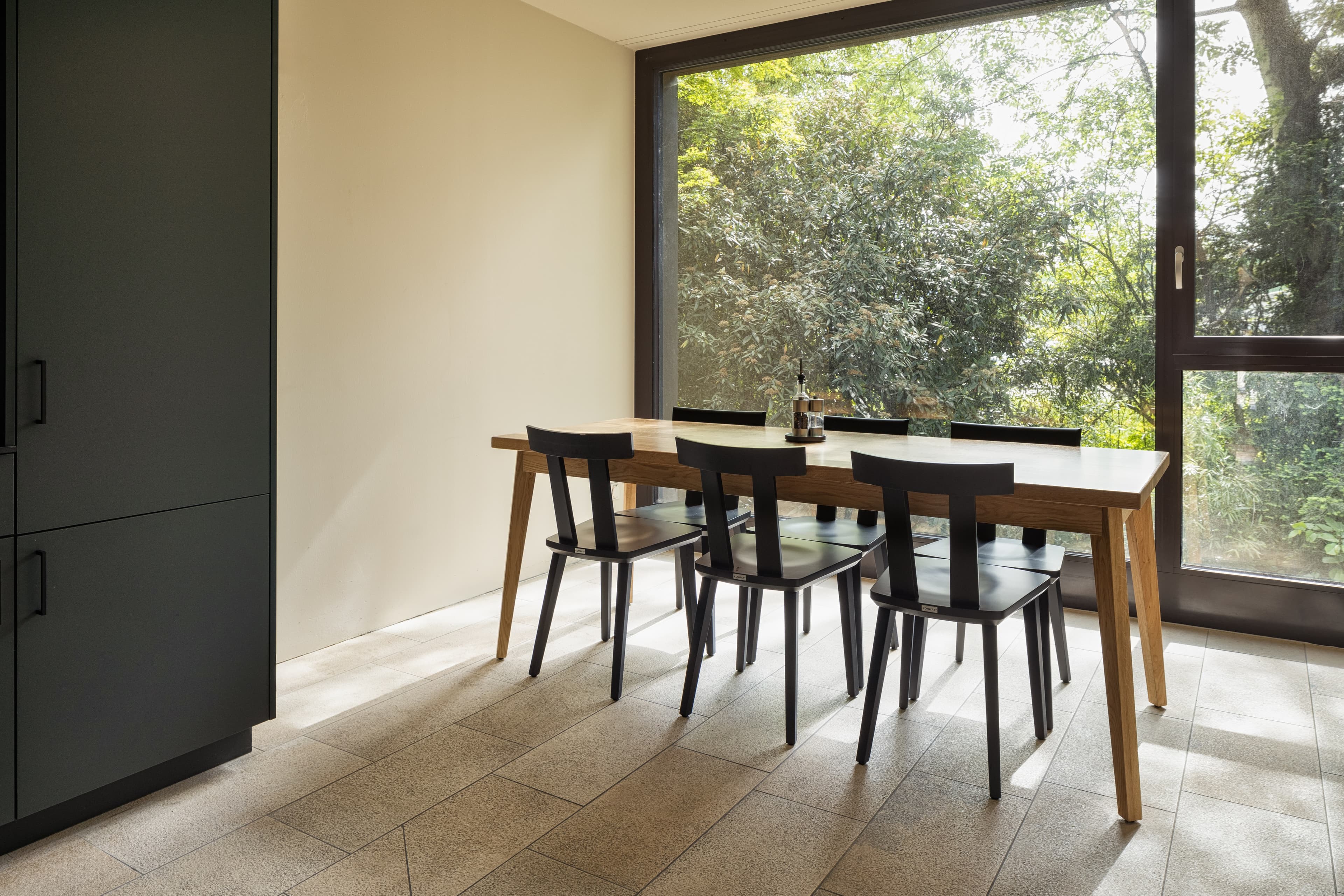 A modern room in the B-Smart Hotel Widnau with a large wooden table, four black chairs, and a window overlooking a green garden.