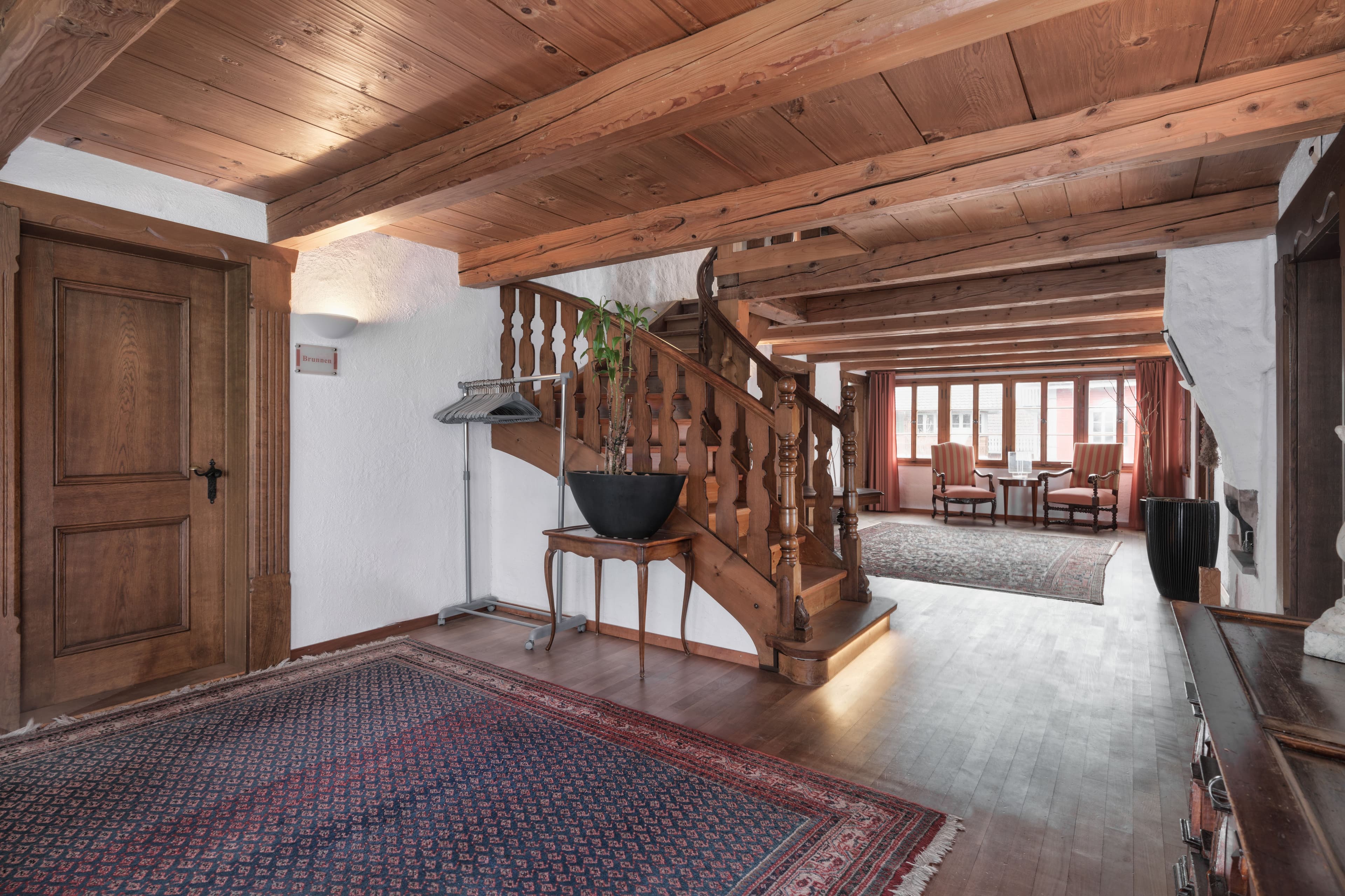 A cozy hotel lobby with a wooden beam ceiling, carpets, a wooden staircase, and a seating area with large windows in the Hotel Kreuz.