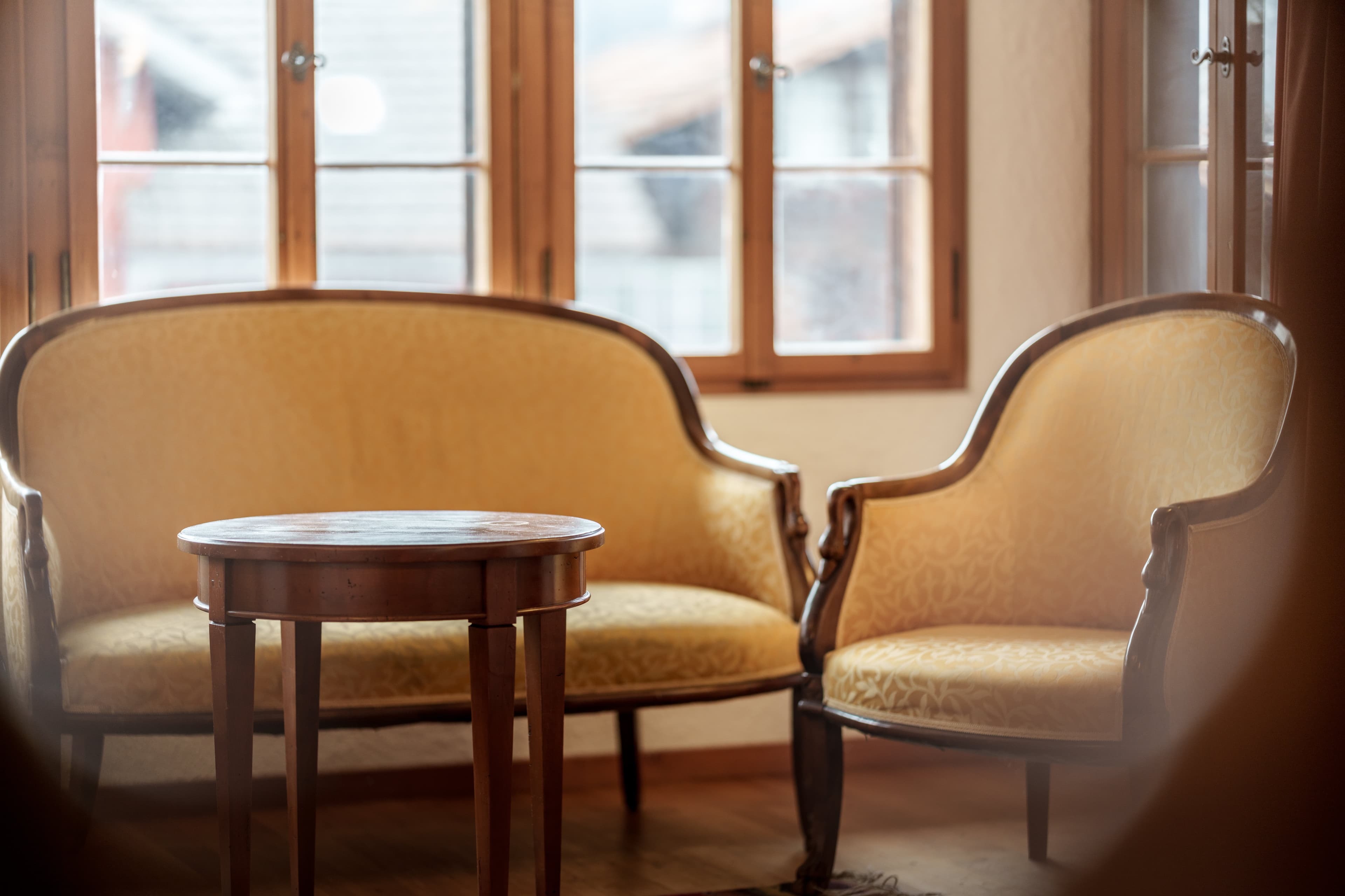 An elegant seating area with a round wooden table, a cushioned sofa, and an armchair in front of large windows at Hotel Kreuz.